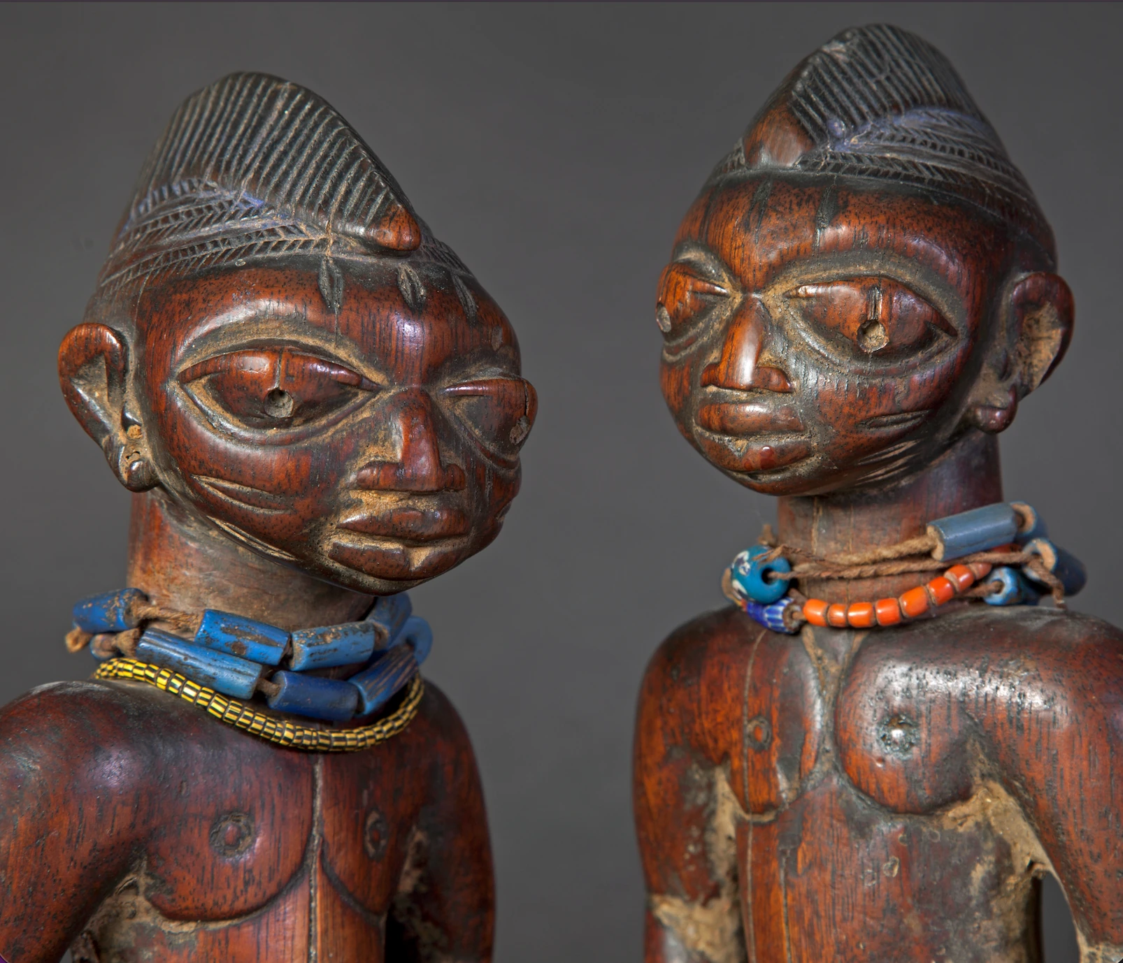 Detalle de los rostros de dos figuras Ibeji de madera, con peinados tallados y collares de cuentas