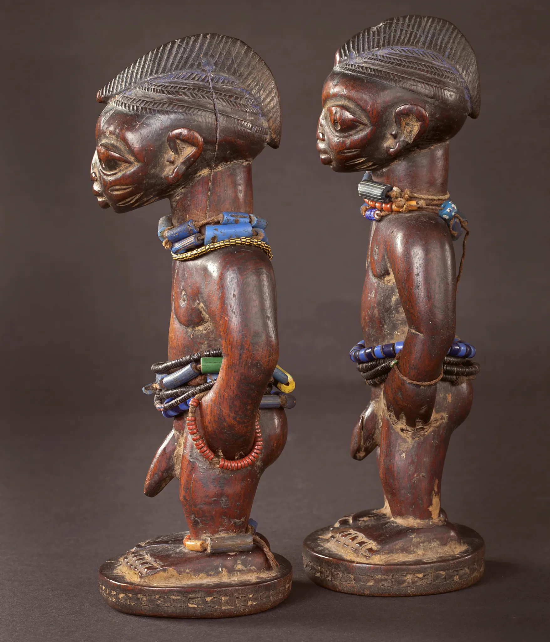 Dos figuras Ibeji vistas de perfil, con peinados tallados y adornos de cuentas
